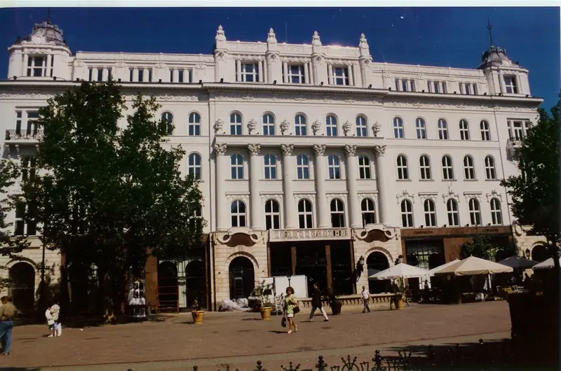 Helle, reich gegliederte Fassade eines großen historischen Gebäudes an einem Platz mit Bäumen und Sonnenschirmen